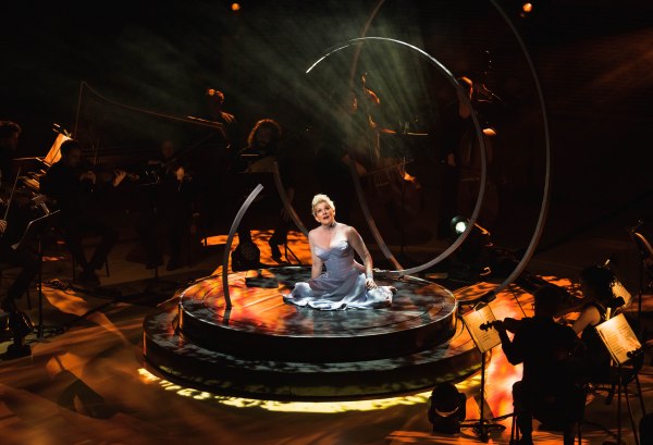 DiDonato sits in a baby blue dress on a black platform illuminated by yellow and orange light. She sings, surrounded by a dark orchestra