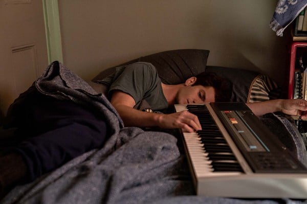 A man laying in bed, next to a keyboard that he places a single hand on.