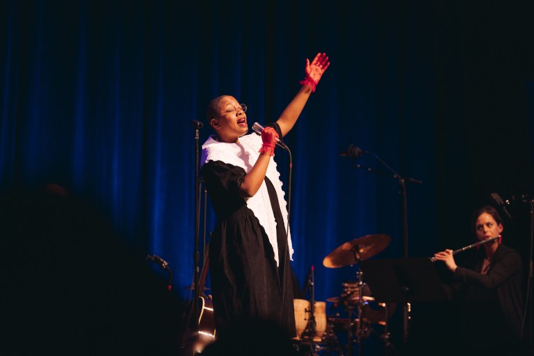 a person with a shaved head and brown skin sings into a microphone, holding one arm up above their head. they wear a black robe with a white piece covering the chest and shoulders