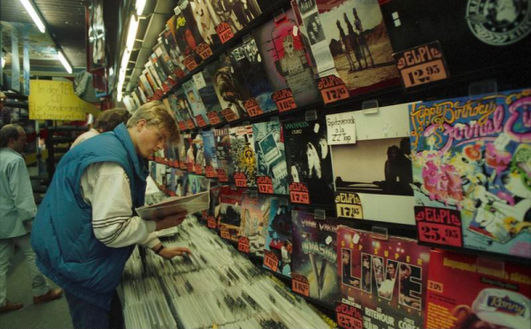 A blond man in a blue vest browses a wall of records in a slightly dim fluorescent light.