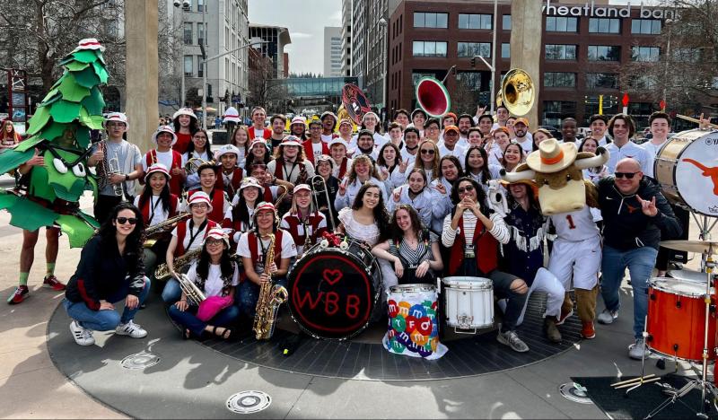 A photo of three dozen smiling students posing for the camera in the middle of a plaza. Several are holding instruments. Rachael Vega is crouched in the front on the left, alongside a student dressed as the Stanford tree.