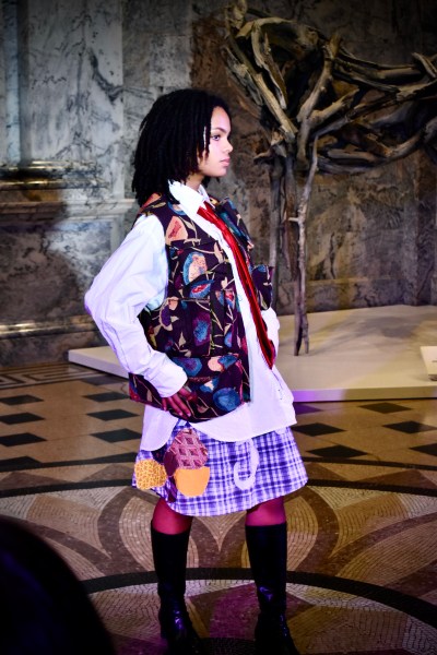 Student model poses at the end of the runway wearing a colorful vest and skirt with a white button-up and boots.