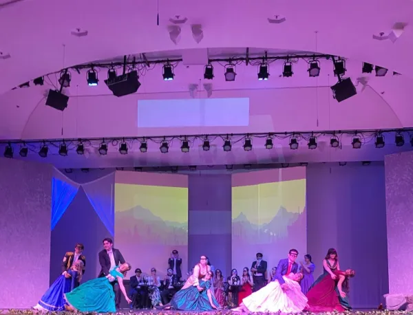 In purple and blue stage lighting, the cast of "Die Fledermaus" dance in ball gowns and tuxedos.
