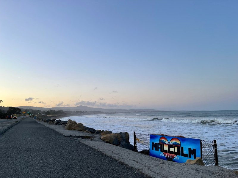 The sun rises over Half Moon Bay. On the left there is an empty road and on the right there is the ocean. In the foreground there is a sign reading "Malcolm: show up and be kind."