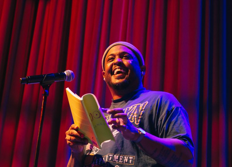 Danez Smith read from their poetry collection with a broad smile on his face in front of deep red theatre curtains in the background. They hold a yellow book and speak into a black microphone in front of them.