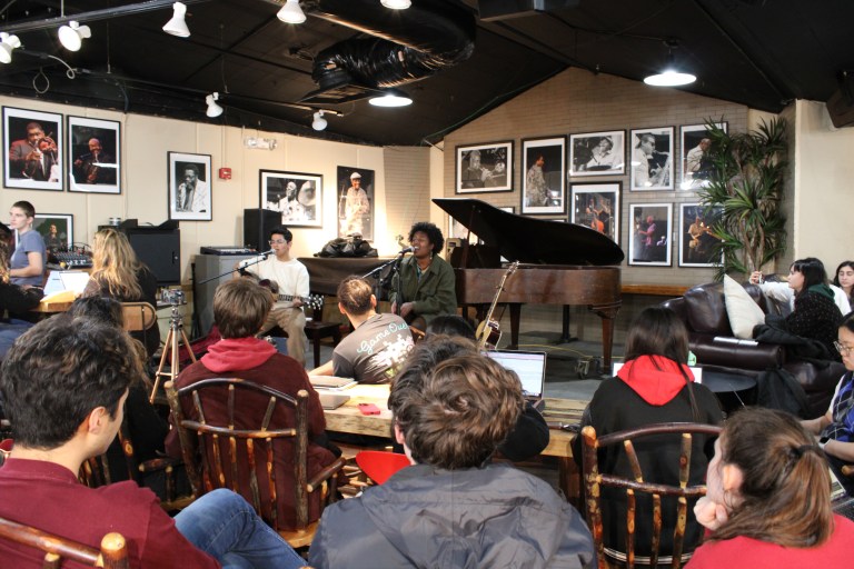 two people sit on a stage singing into microphones. the person on the left has light skin and short brown hair, and strums a guitar. the person on the right has brown skin and short curly black hair, and seems to be clapping to a beat