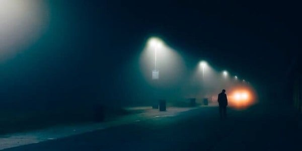 A man walks listlessly along a dark street lit intermittently by streetlights.