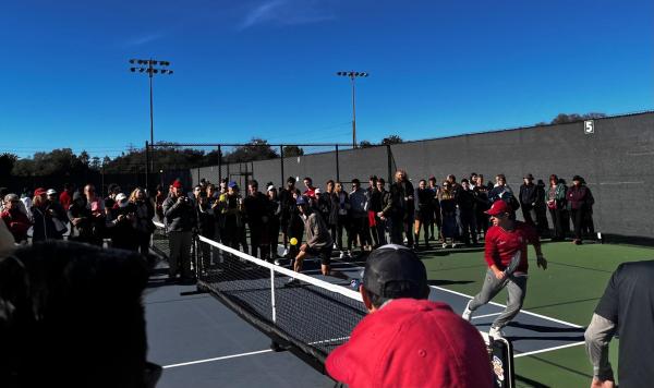 A large crowd stands around a Pickball court as a player returns a shot.