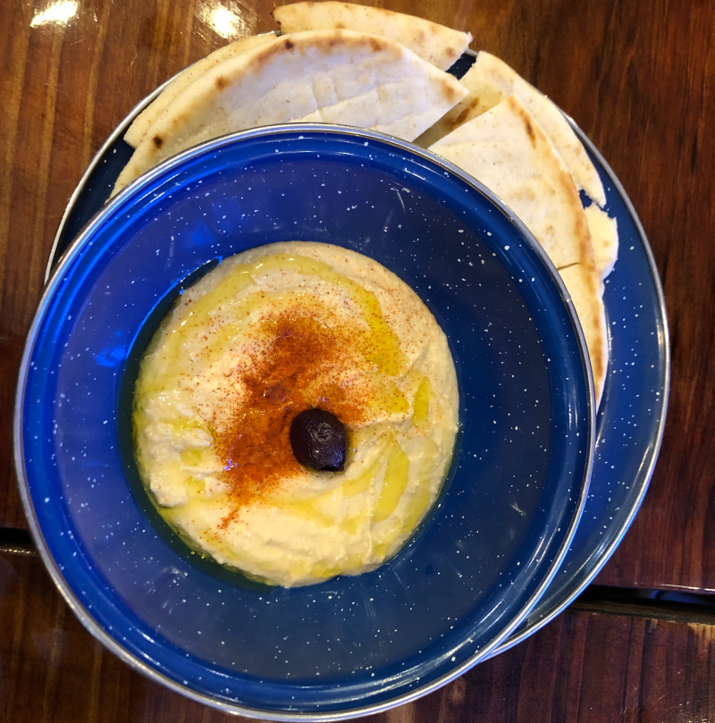 A speckled blue bowl holds hummus with olive oil, paprika and an olive.