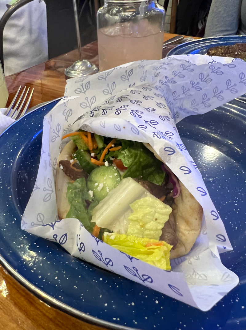 A lamb pita wrapped in white paper with blue details sits on a speckled blue plate.