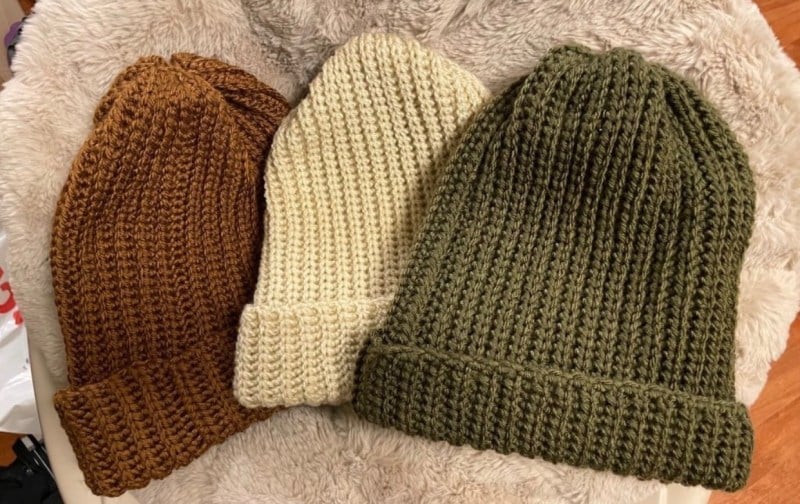 Three crochet beanies in brown, white and green lay against a rug. 