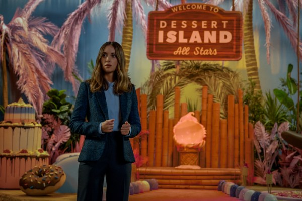 a photo of a young white person with medium-length straight hair, standing on the set of a movie in front of a sign that reads, "Dessert Island: All Stars"