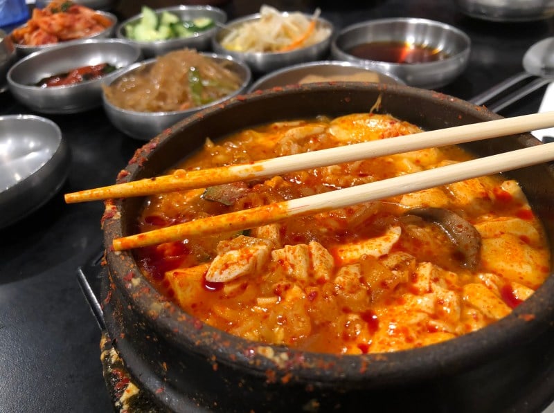 Tofu soup steams in a stone bowl with chopsticks resting on top of the bowl.