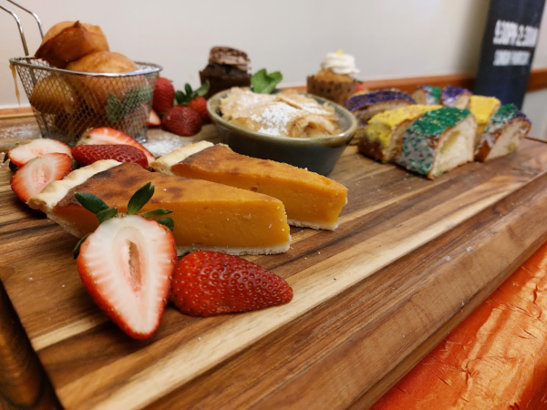 a photo of a wooden cutting board with fruit, sweet potato pie, beignets, and colorfully sprinkled king cake