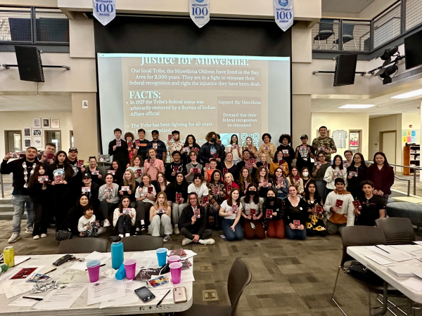About 70 high school students and members of the Muwekma Ohlone Tribe pose for a photo, with the screen behind them reading "Justice for Mukwema"