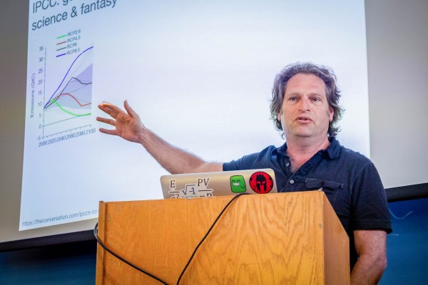 Adam Aron stands at a podium pointing to a slide titled "science & fantasy," which contains line graph with projections of emissions over time