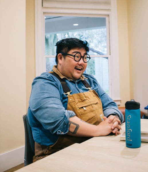 Hieu Minh Nguyen talks with a smile. He is waring a yellow ochre colored overalls and a blue button up underneath . He also has round black glasses on