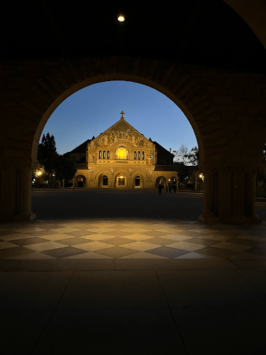 A lit up Memorial Church framed in the center of an arch. It is almost night.