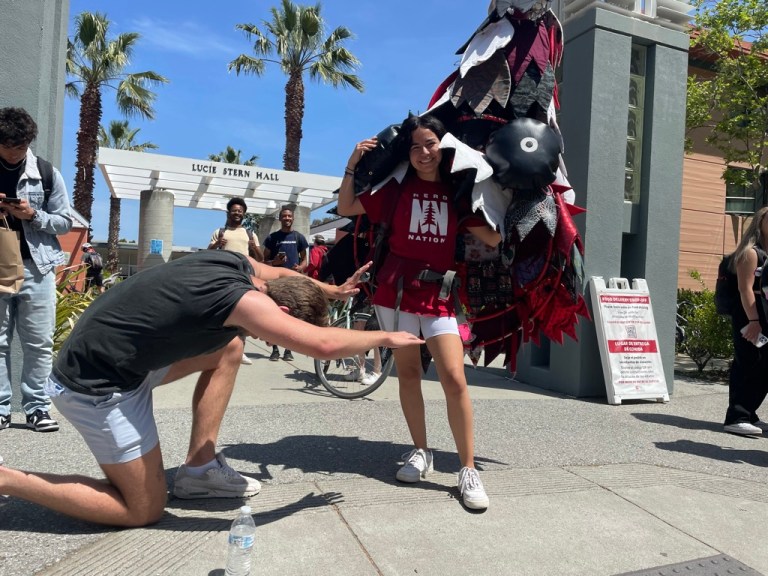 a photo of a person wearing a cartoonish costume of a tree with black and white leaves, wide eyes and a big mouth. there is another person in a t-shirt and shorts kneeling before them.