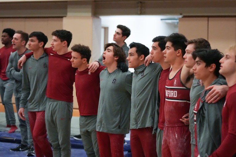 Gymnasts stand in a line with their arms around one another, watching at a gymnastics meet.