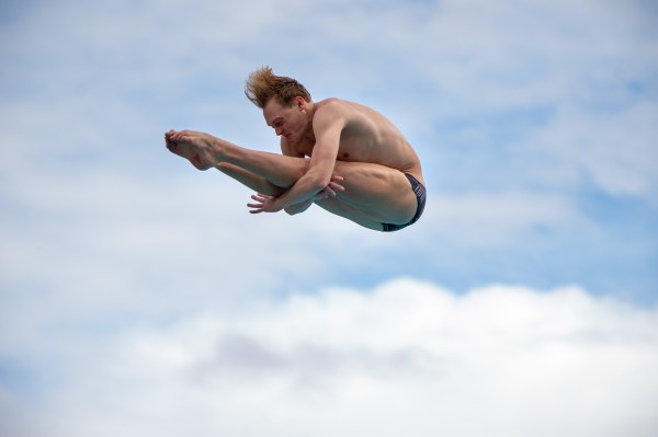 Jack Ryan holds a pike position mid-air during a diving competition, with the sky and clouds in the background.