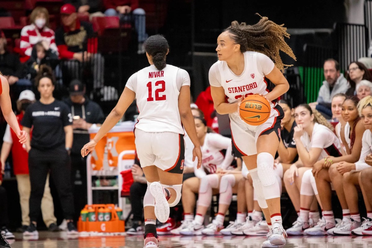 An unedited action shot of Stanford Women's Basketball player #30 Hayley Jones carrying a basketball and her team member #12 Kiki Iriafen passes her. Fans, a gatorade stand and the women's basketball team stands behind her.
