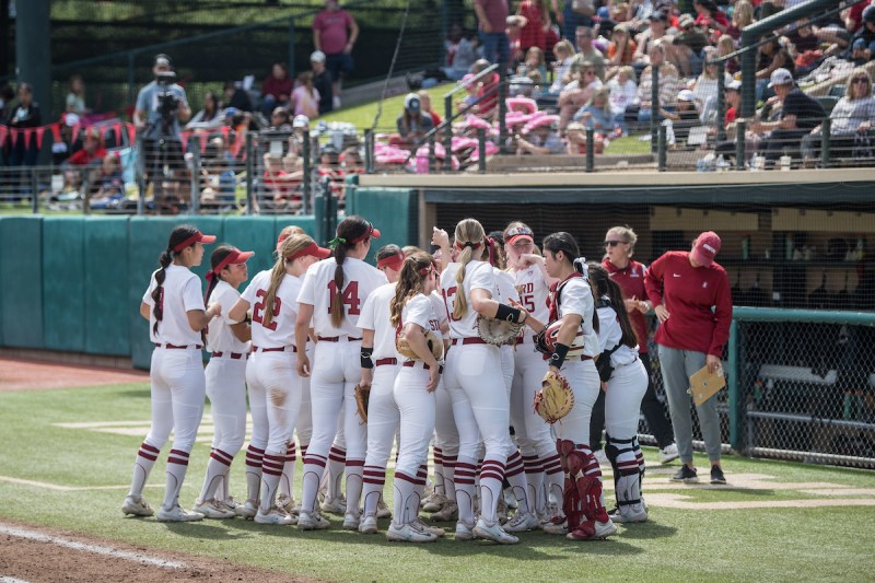 A group of Stanford softball players gathers during a game as the crowd watches on