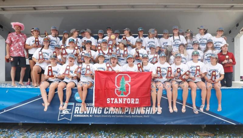 Athletes pose in three rows with a Stanford flag and hold their trophies after winning the NCAA Women's Rowing Championship.