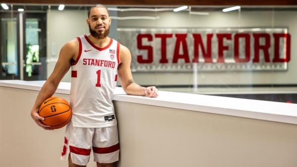 A man holding a basketball in athletic clothes standing next to a Stanford sign.