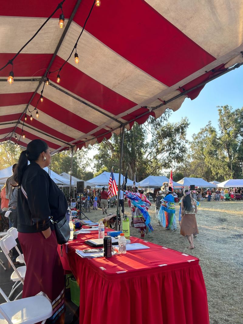 A woman stands at her booth at the Stanford Powwow watching the dancers in Eucalyptus Grove. 