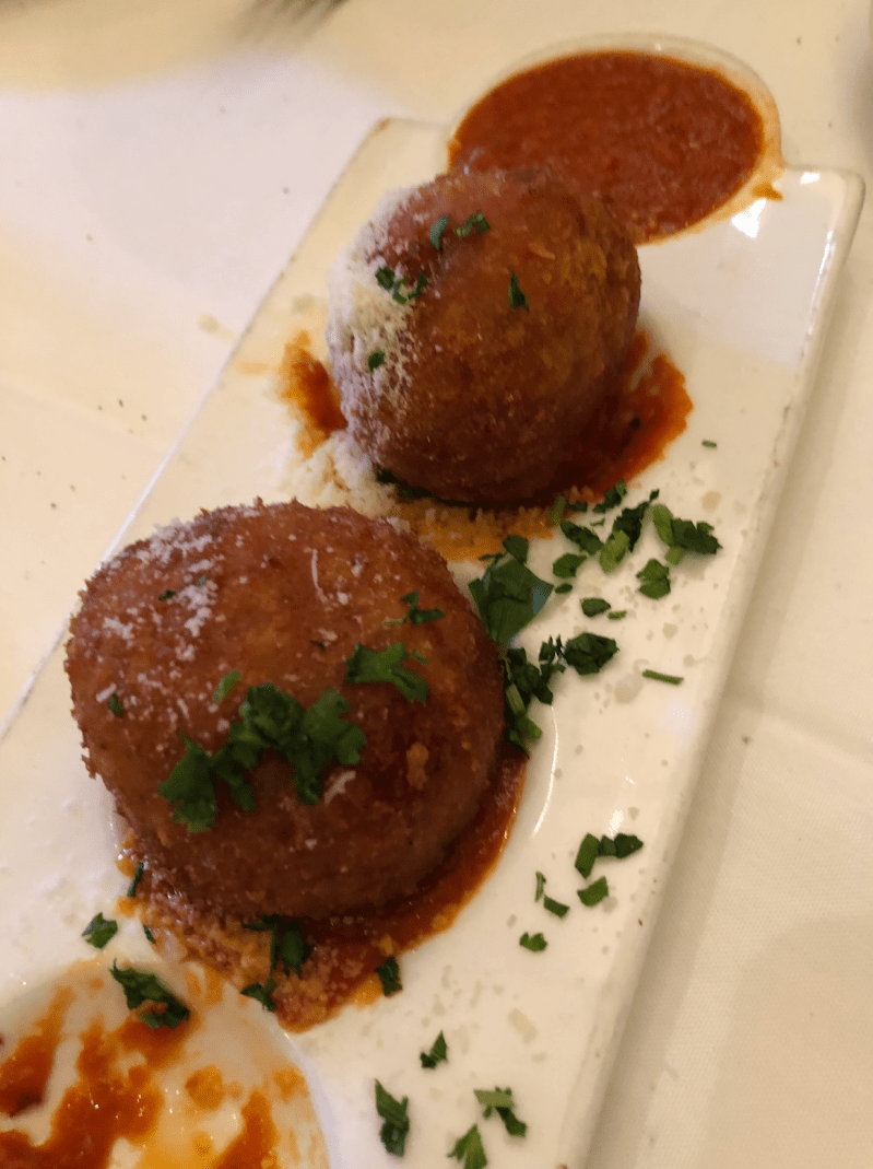 Two arancine balls sit in a small white dish with sides of tomato sauce and cheese.