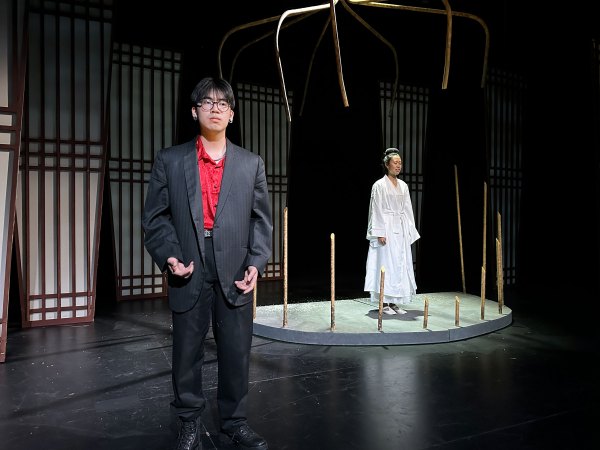 A boy in a red shirt and black blazer faces the audience of TAPS' performance of "Snow in Midsummer." A girl in a white dress in a large, cage-like structure stands behind him.