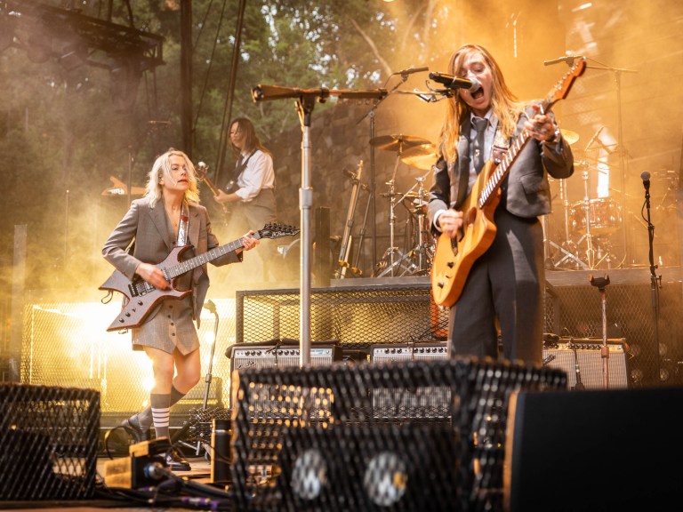 a photo of two people with light skin and blonde (left) and brown (right) hair singing and playing guitars on a stage. other musicians can be seen in the background