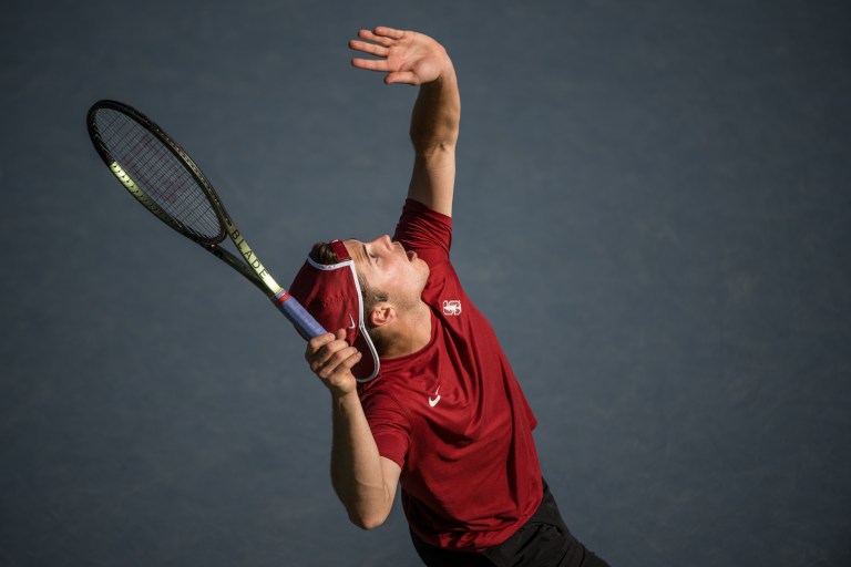 Tennis player Arthur Fery bends on arm back behind his head as he prepares to serve the ball during a match.