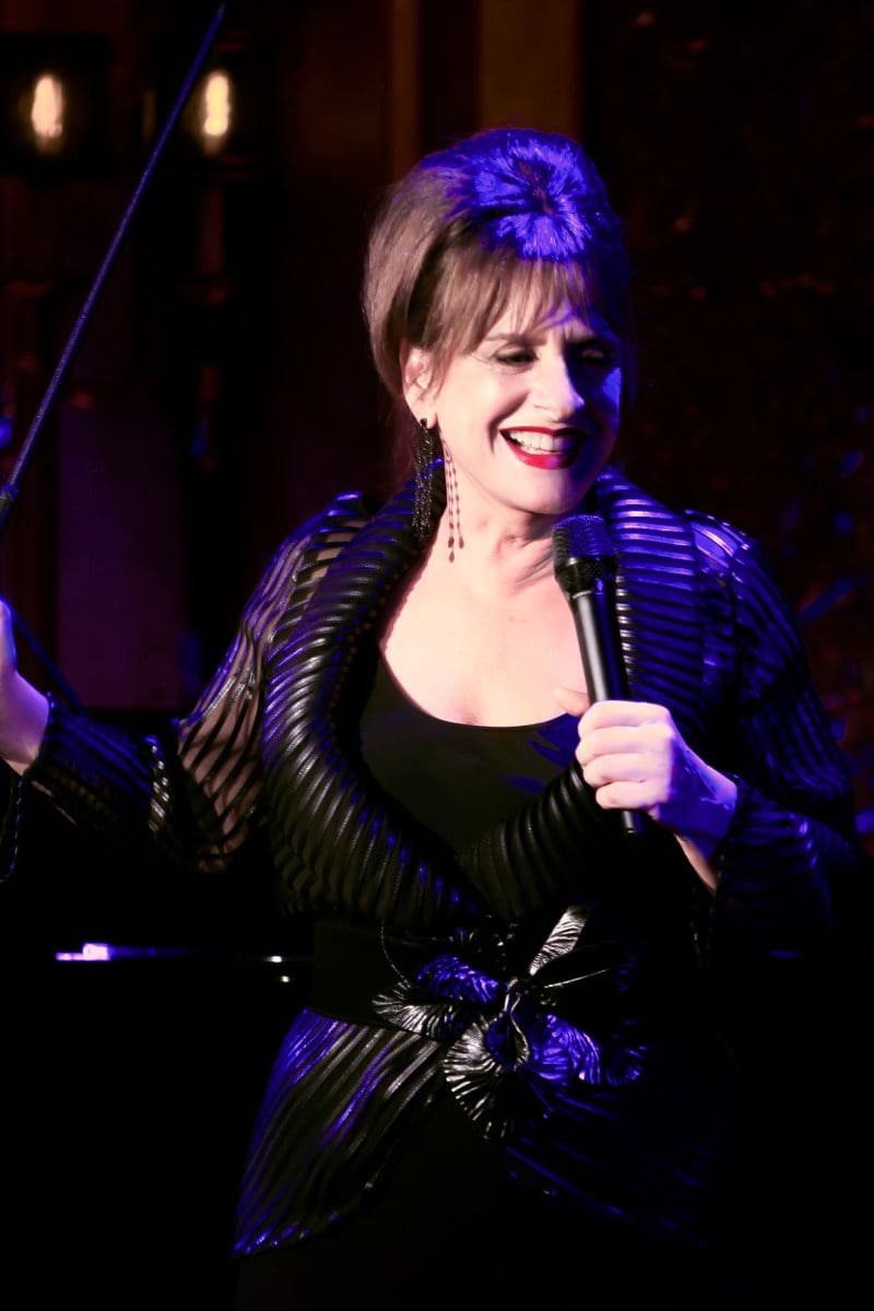 a photo of a feminine person with light skin and brown hair in an updo with bangs. the person is holding a microphone while illuminated by a spotlight