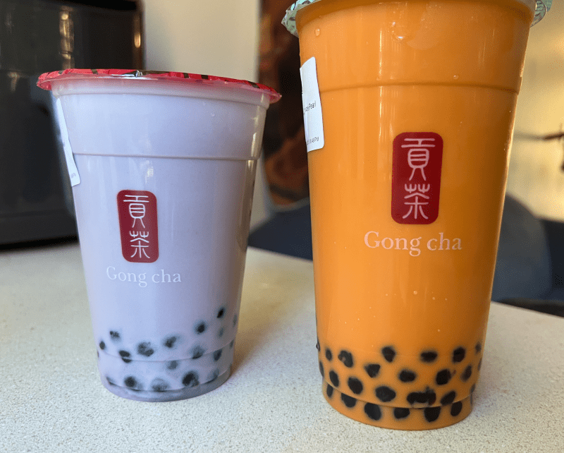 A small purple ube latte and large orange Thai tea drink from Gong Cha sit on a desk.