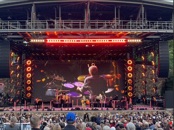 a photo of a stage with several instruments and musicians. video footage of the drummer is being shown on a large screen behind the performers. the audience can be seen in front of the stage.