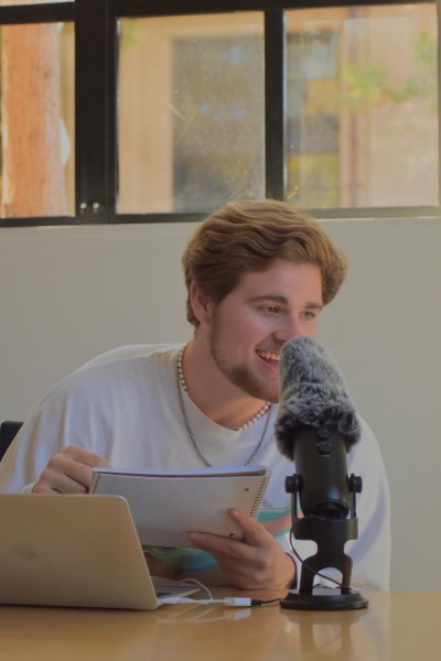 a photo of a person with light skin and short brown hair sitting at a desk in front of a laptop, holding a notebook, and speaking into a microphone