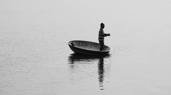 Black and white photo. A man fishes from a small boat in the middle of a lake.