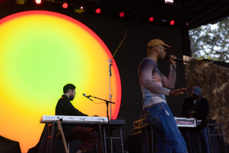 Toro y Moi performs on stage at Frost Amphitheater wearing a brown cap. 