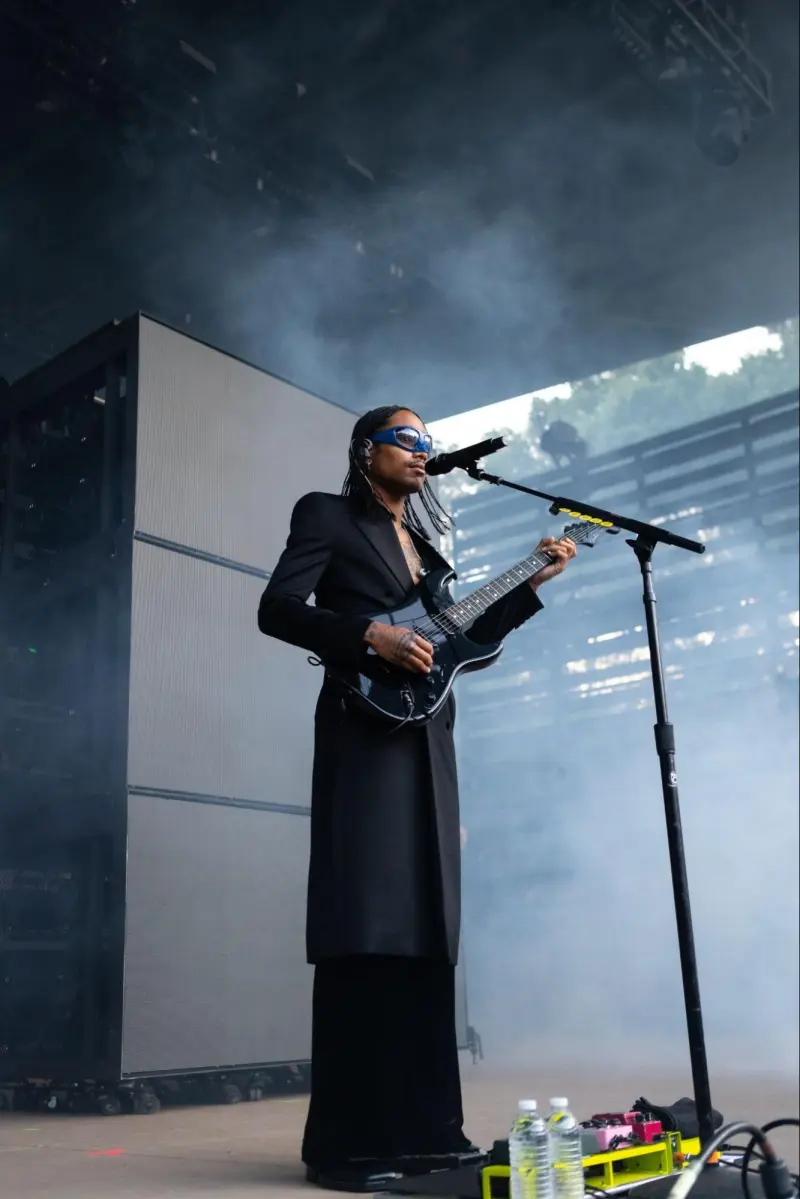 Steve Lacy stands on Frost Amphitheater's stage wearing a long black blazer and pants with an electric guitar.