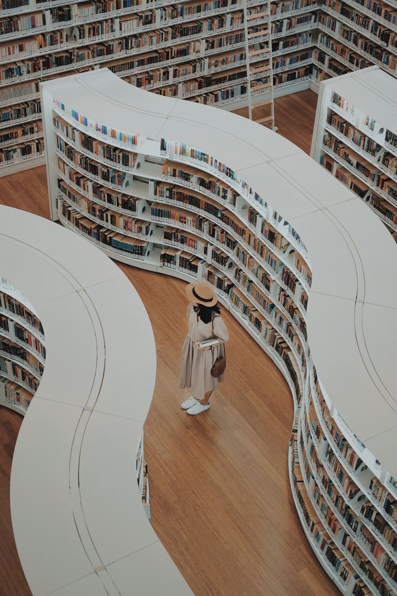 A woman in a white hat stands between white curving bookshelves full of colorful books.