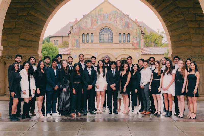 a group of 31 people dressed formally in suits and dresses stand beneath an arch in Stanford's Main Quad, with the pointed roof and painted murals of Memorial Church visible in the background