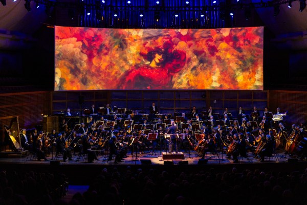 A photo of a symphony orchestra in the dark. In the background is a screen displaying an image of a woman in red.