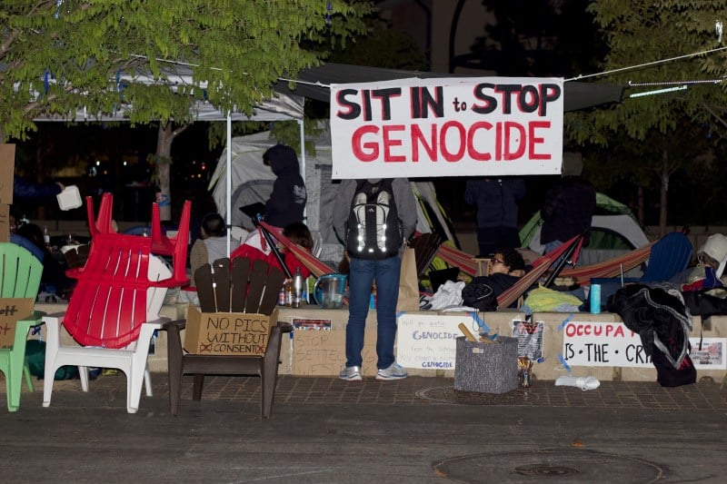 Tents and chairs set up on White Plaza at night, with protestors camping. A poster hanging reads "Sit In to Stop Genocide"