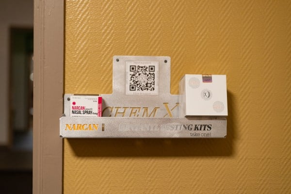 Shelf of Narcan and fentanyl test strips hanging on a door, along with a QR code to scan for more info.