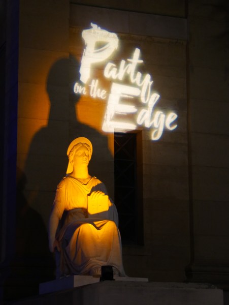 A dark room lit by a lamp shaped like a Greek statue. LED lights display "Party on the Edge."