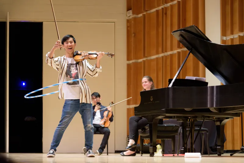 Sophie Druml perches on a piano bench and Brett Yang of TwoSet Violin uses his phone to capture the scene of TwoSet Eddy Chen brandishing his bow while playing the violin and hula hooping onstage.