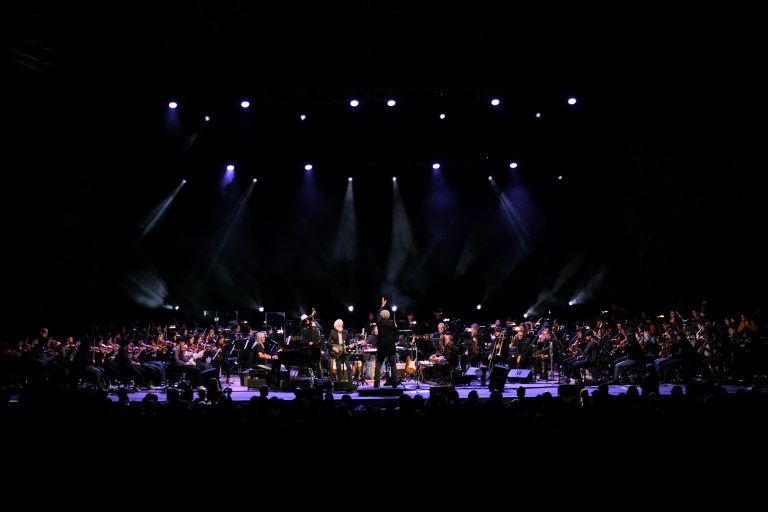 A five-person rock band is center stage, surrounded by a 115-person orchestra dressed in black. The scene is dark and dramatic.