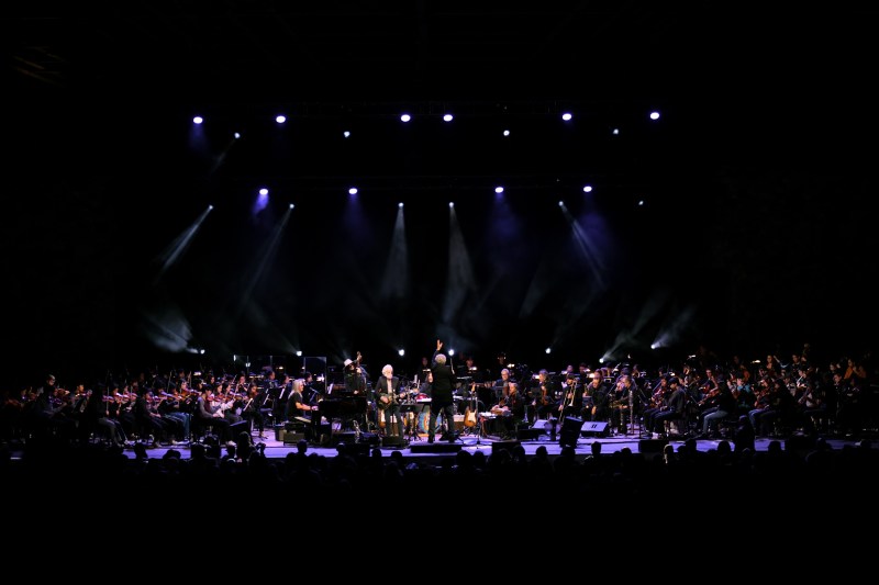 A five-person rock band is center stage, surrounded by a 115-person orchestra dressed in black. The scene is dark and dramatic.